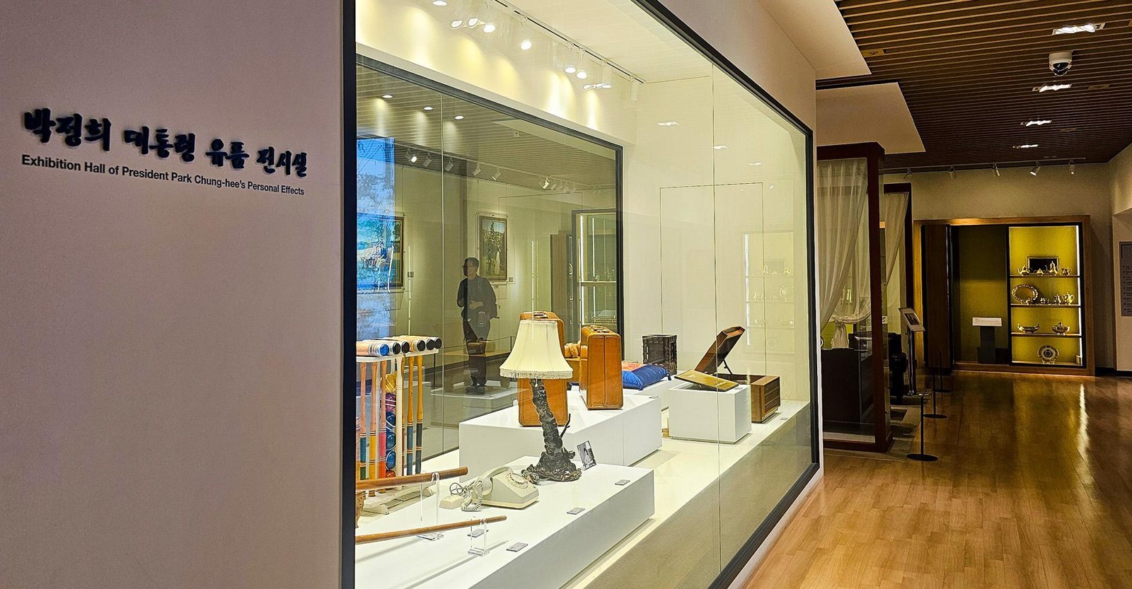 유품 전시실 — 왼쪽 벽 글자 “박정희 대통령 유품 전시실 / Exhibition Hall of President Park Chung-hee’s Personal Effects”, 유리 진열장 안 크로켓 채·스탠드·다이얼 전화기·가죽 가방·방석, 오른쪽 나무 바닥 복도와 금빛 진열장